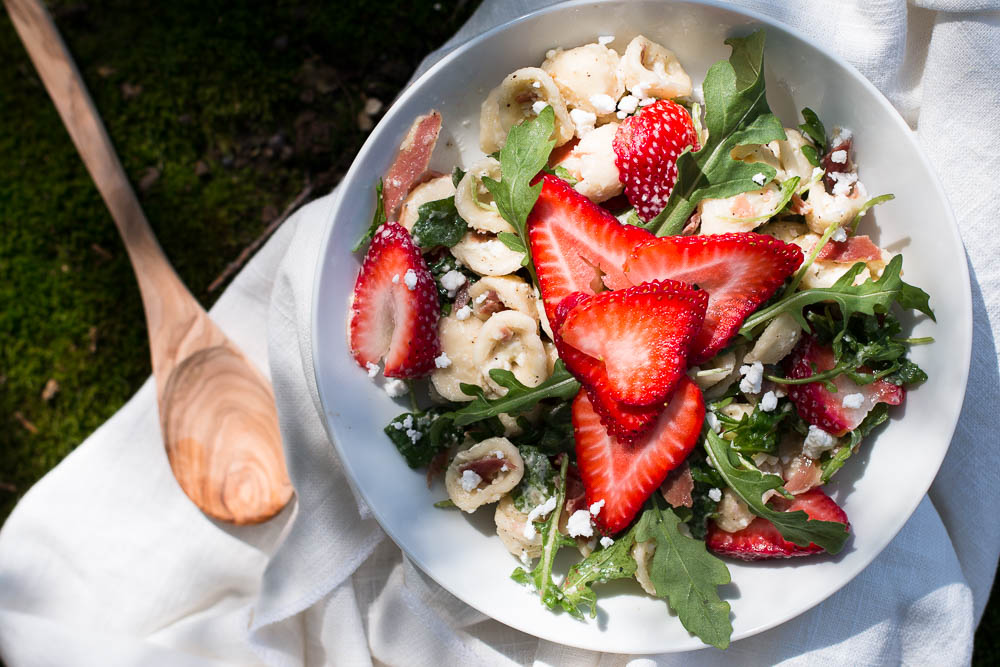 Overhead shot of orecchiette pasta salad with arugula, feta, and strawberries in a shallow white bowl next to a wooden spoon.