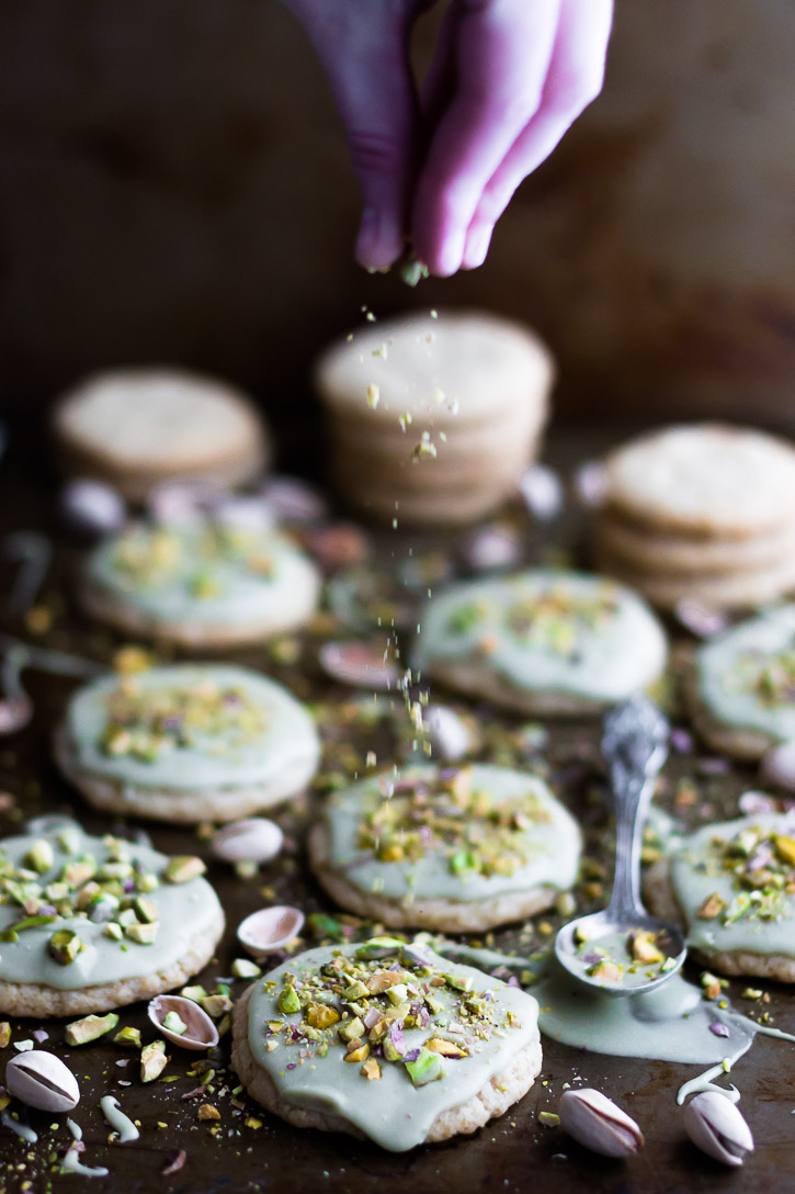 Vertical side shot showing a hand sprinkling frosted cookies with ground pistachios.