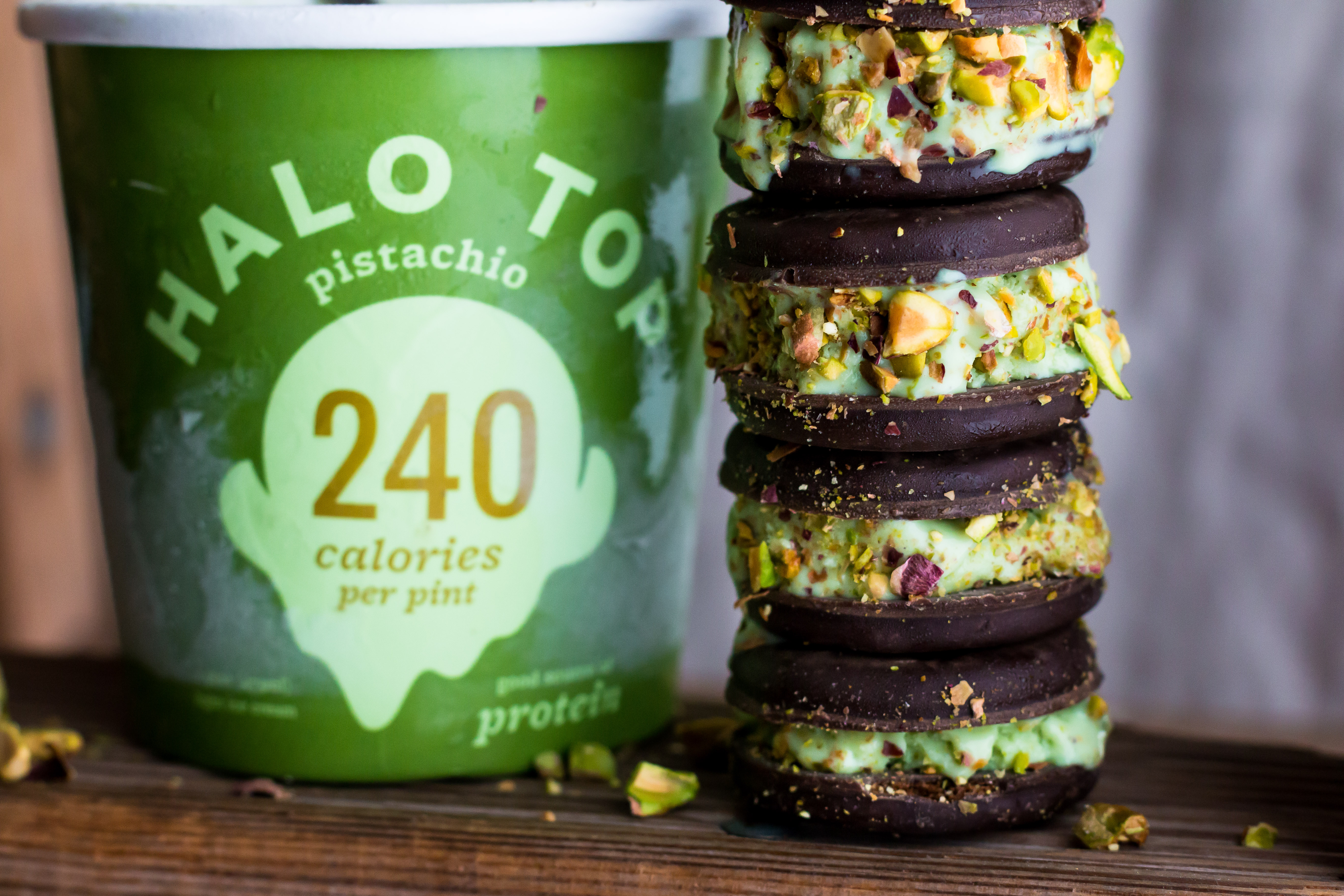A close up horizontal side shot of four stacked pistachio ice cream sandwiches next to a pint of Halo Top ice cream with a spoon in it.