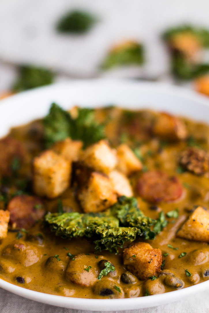 Close up vertical view of black beans, croutons, and kale chips in the Creamy Red Pepper and Andouille Sausage Soup.