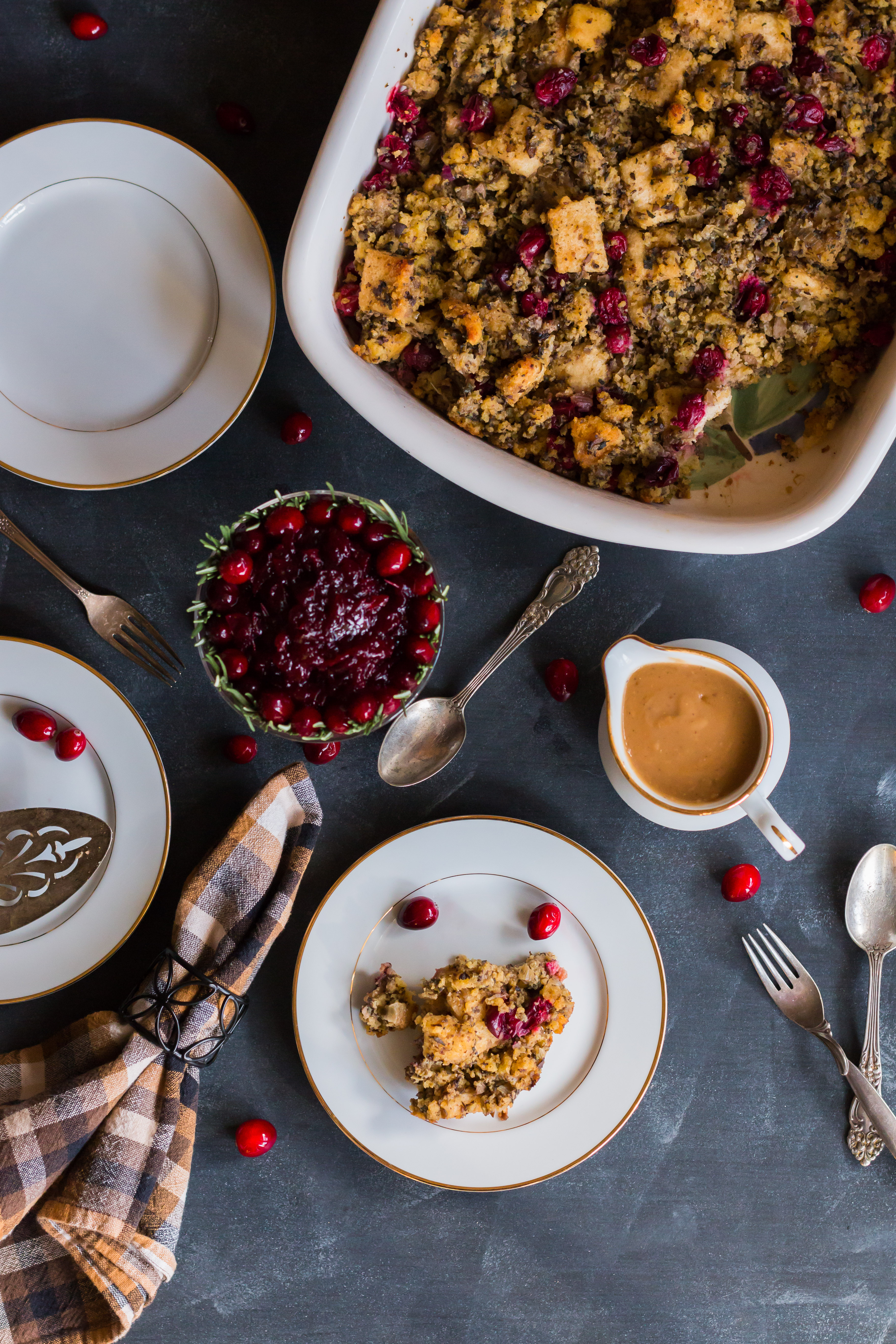 Overhead shot of stuffing in a rectangular casserole dish, cranberry sauce in a round serving dish lined with cranberries and rosemary, gravy in a sauce server, three small plates with a serving of stuffing, a napkin, and silverware.