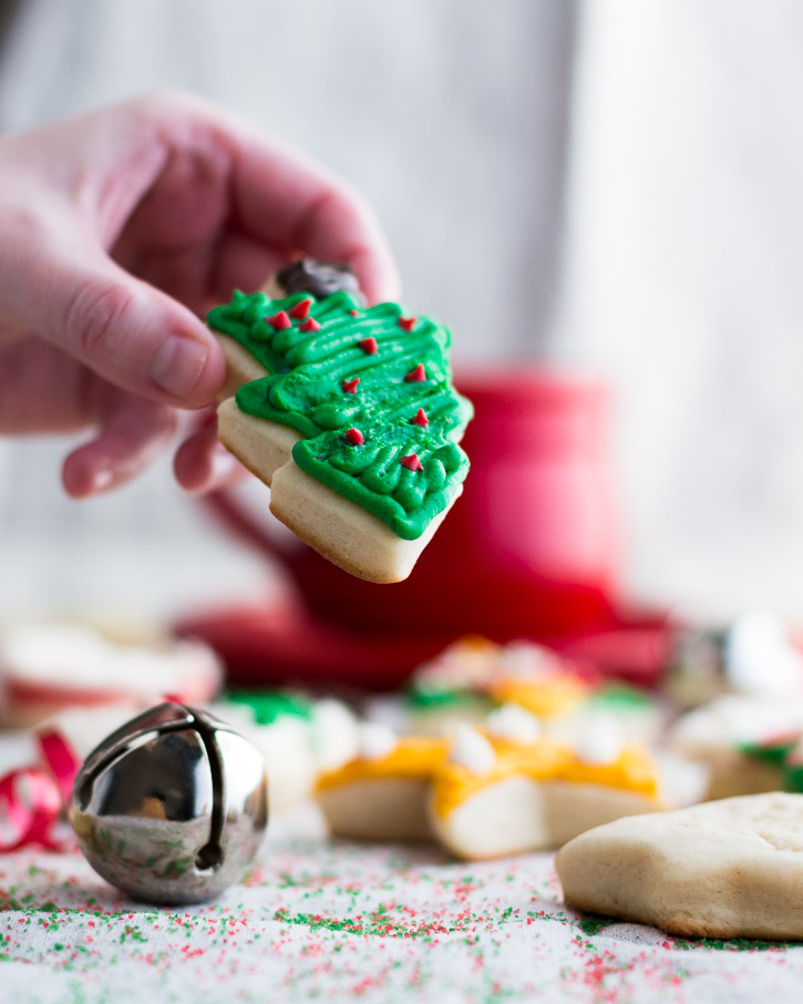 Vertical shot of a hand picking up and lifting a frosted Christmas tree shaped cookie in front of a bell and other cookies.