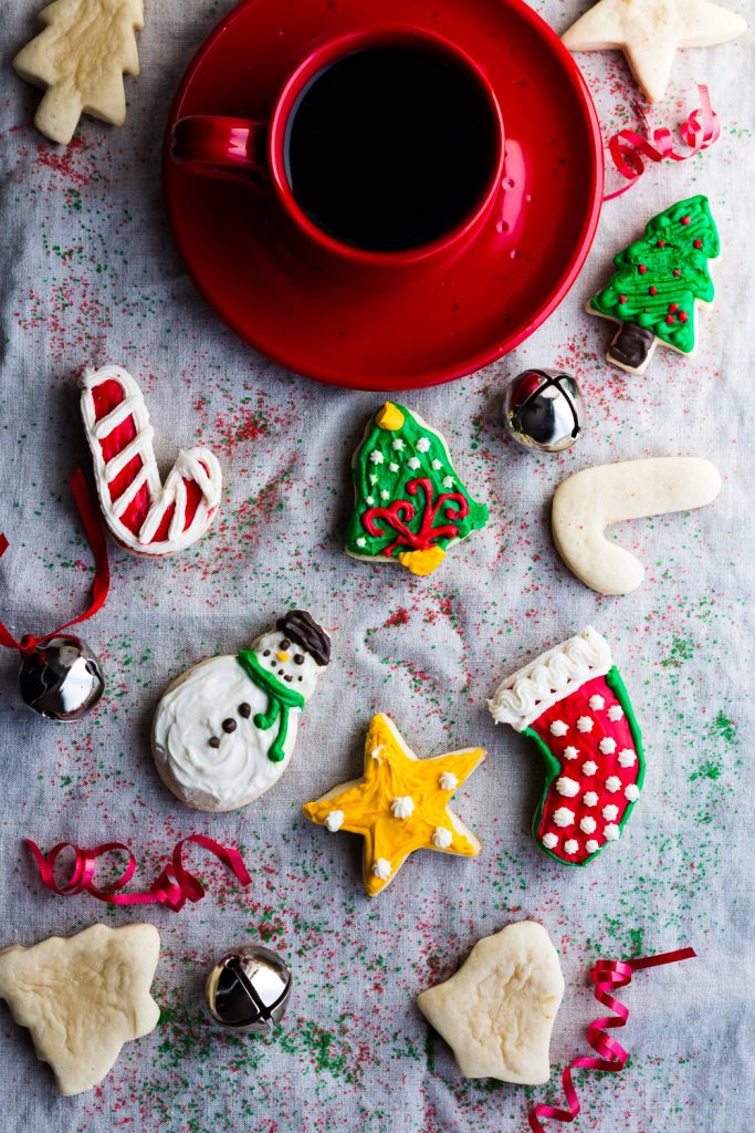 Overhead view of frosted Christmas cookies surrounded by bells, a coffee cup and saucer, and curled ribbon on top of a sprinkle covered clothe back round.
