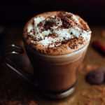 Slightly angled vertical view of glass mug with metal handle filled with hot chocolate, topped with whipped cream and chocolate shavings, and surrounded by cocoa powder, chocolate wafers, and dried chilies.