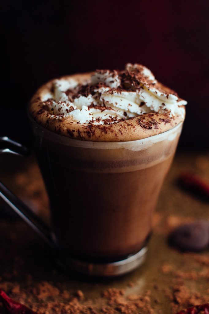 Slightly angled vertical view of glass mug with metal handle filled with hot chocolate, topped with whipped cream and chocolate shavings, and surrounded by cocoa powder, chocolate wafers, and dried chilies.