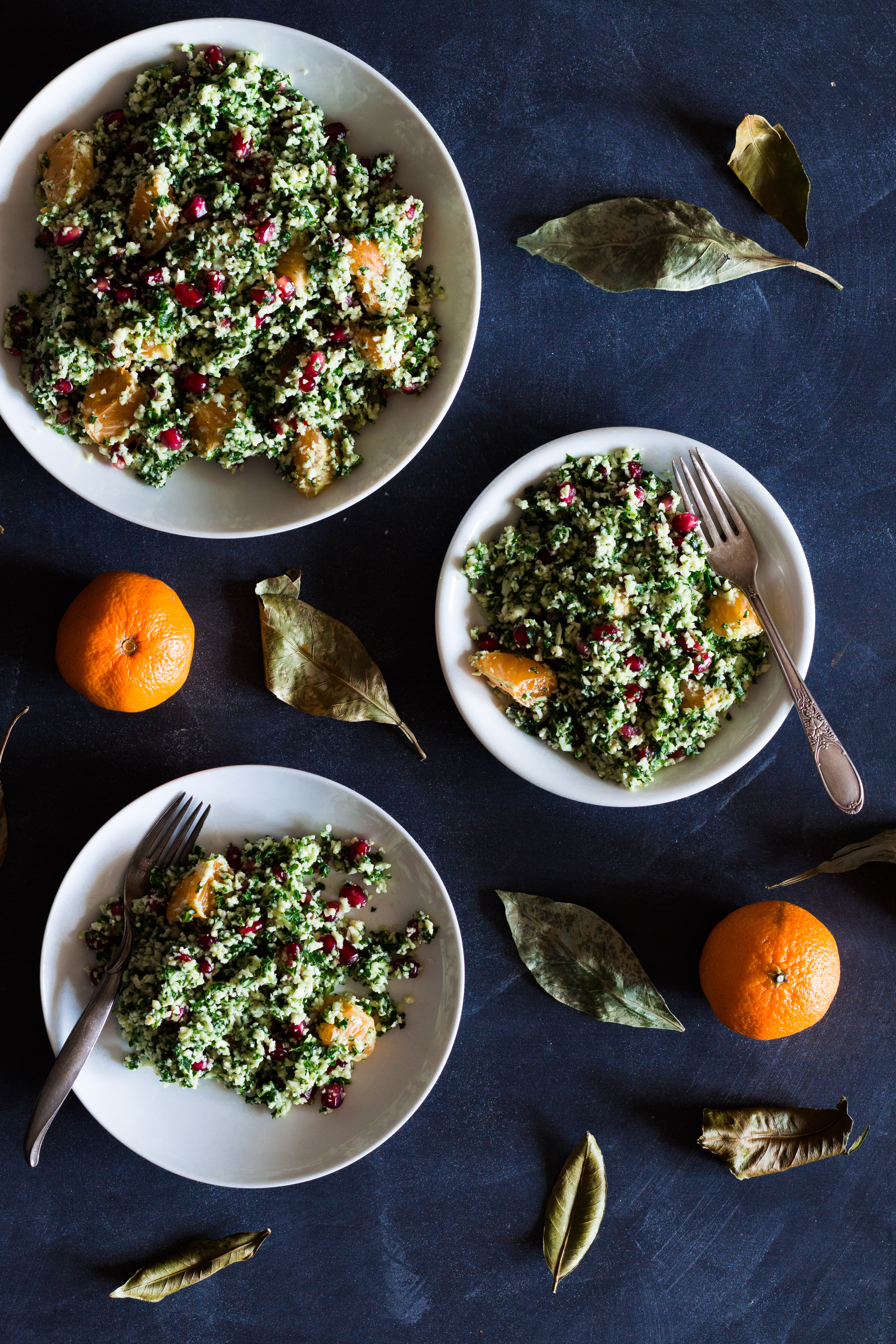 Overhead shot of three circular plates with servings of cauliflower and kale tabbouleh on a dark blue surface and surrounded by tangerines and green leaves.