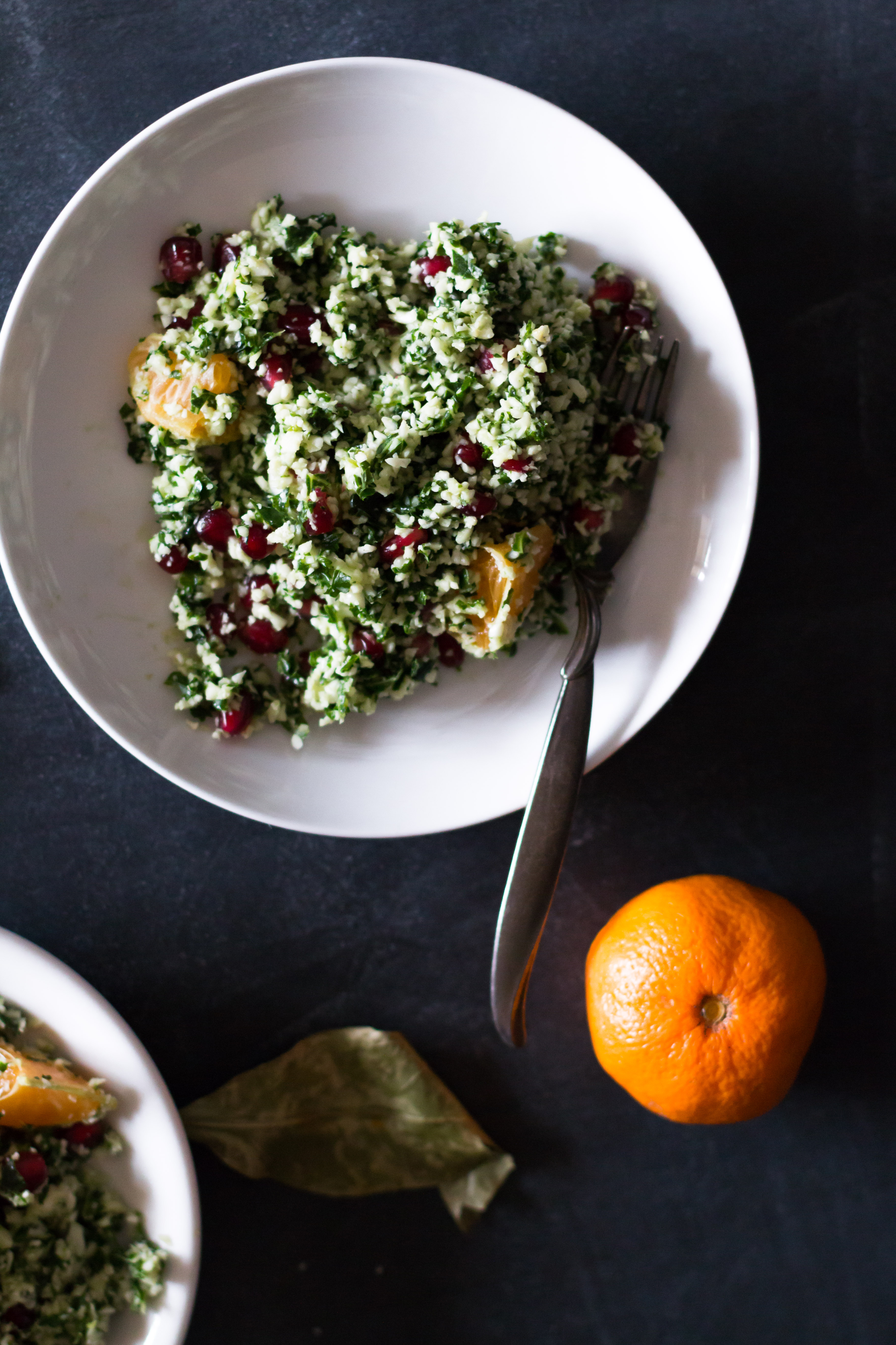 Over head shot of one circular plate filled with cauliflower and kale tabbouleh and a fork on a dark blue surface and surrounded by a tangerine and leaf.