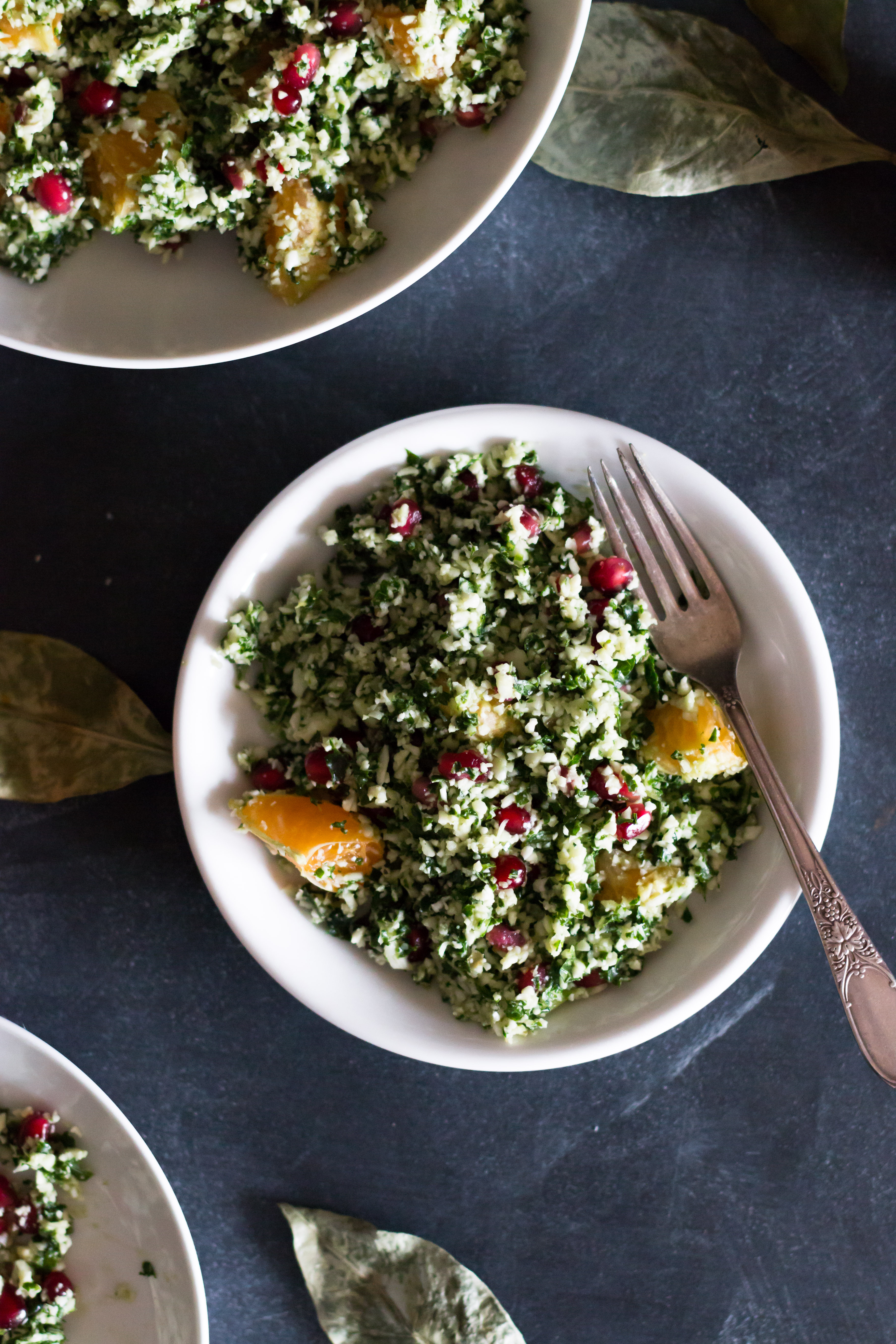 Over head shot of one circular plate filled with cauliflower and kale tabbouleh and a fork on a dark blue surface and surrounded by leaves and parts of other plates.