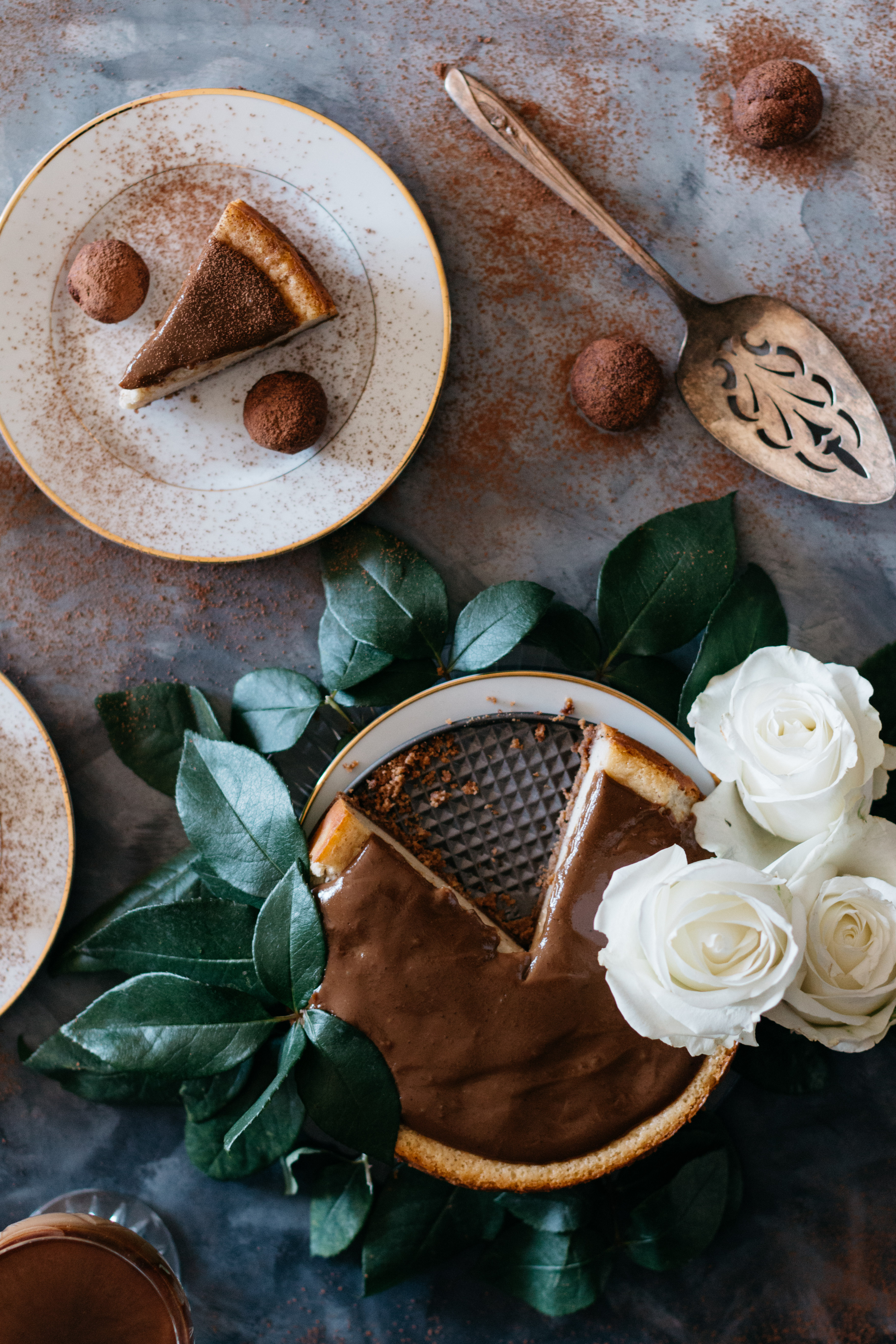 Overhead shot of a cheesecake surrounded by leaves and roses next to a plate with a cheesecake slice, truffles, and a pie server.