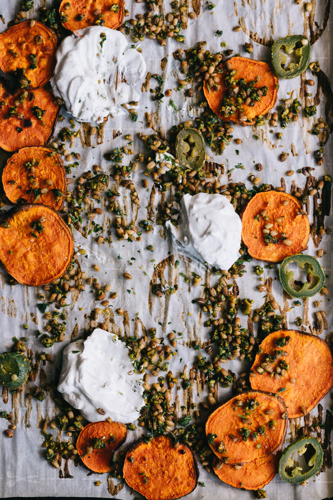 Overhead shot of parchment covered baking sheet that's been used as a serving platter people have eaten off of. There are a few sweet potato crisps leftover, gremolata and barley scattered everywhere, and dollops of dip that have been used.