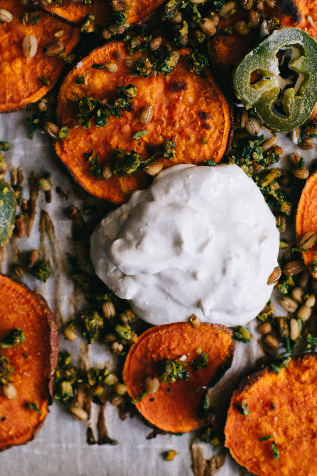 Close up overhead shot of a dollop of sour cream blue cheese dip on a parchment paper covered baking sheet surrounded by sweet potato crisps, gremolata, barley, and jalapeño slices.