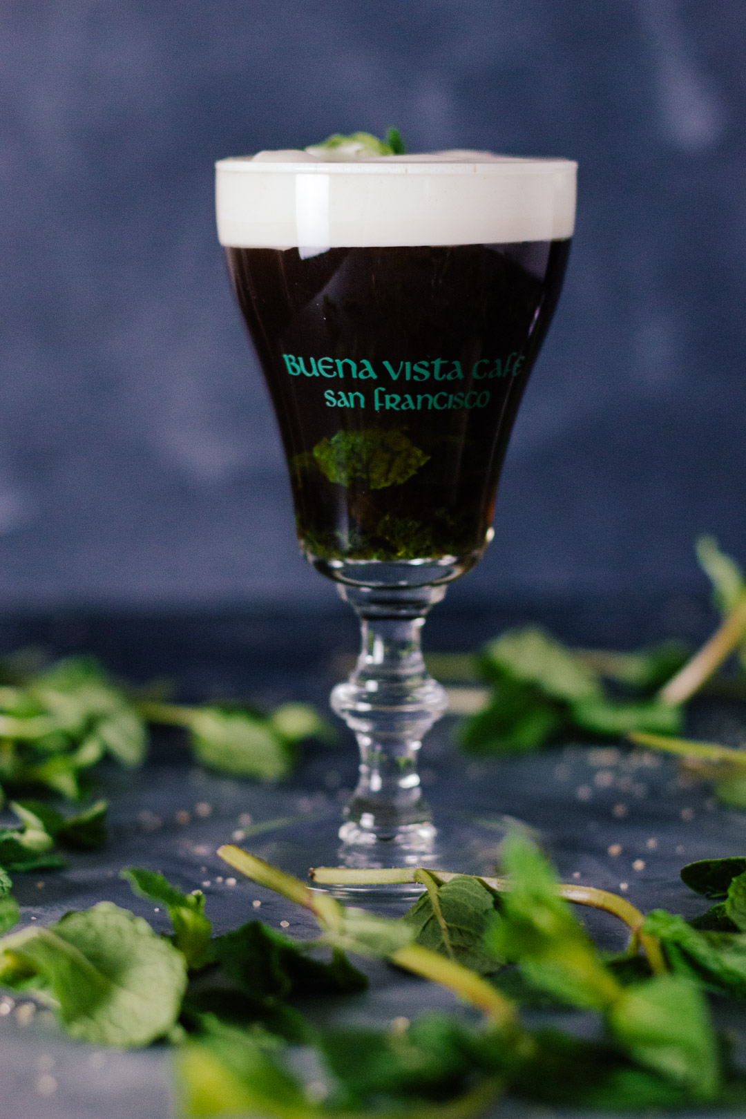 Vertical shot of an Irish Coffee topped with cream in a small glass goblet surrounded my mint leaves and grains of sugar.