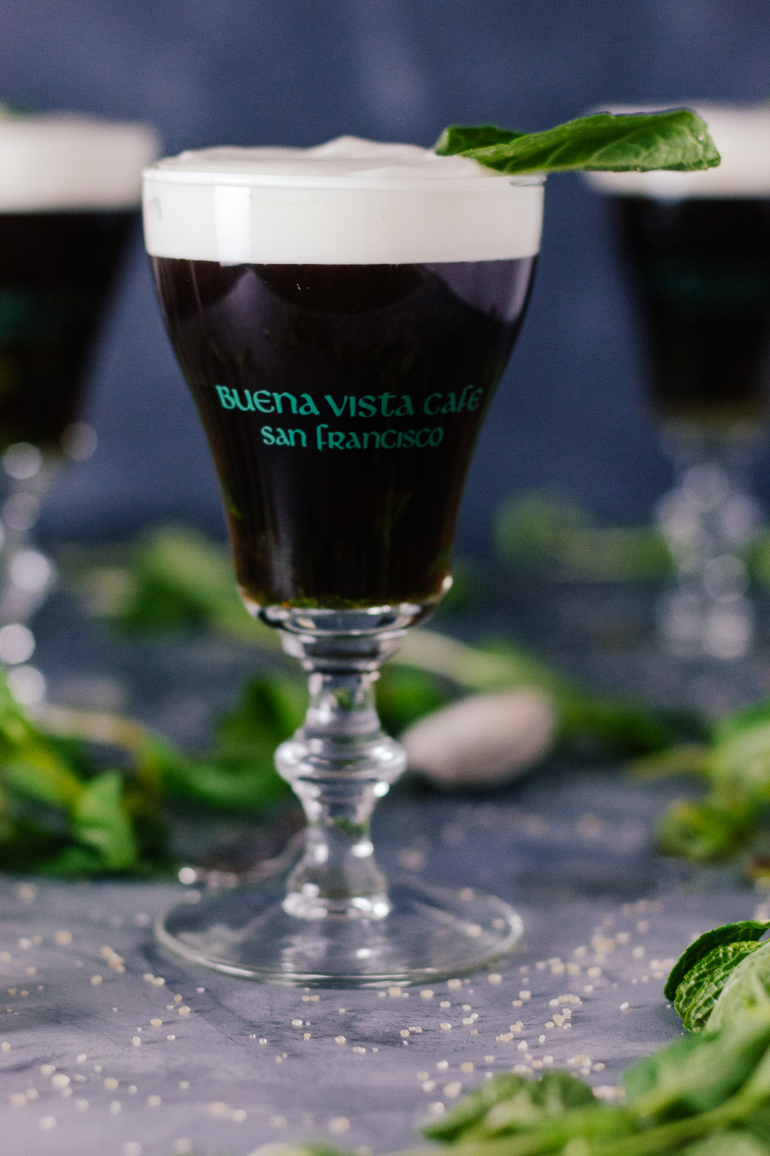 Vertical shot of an Irish Coffee topped with cream in a small glass goblet surrounded my mint leaves, grains of sugar, and other Irish Coffees.
