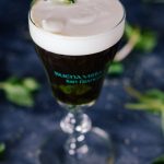 Vertical, slightly angled shot of an Irish Coffee topped with cream in a small glass goblet surrounded my mint leaves and grains of sugar.