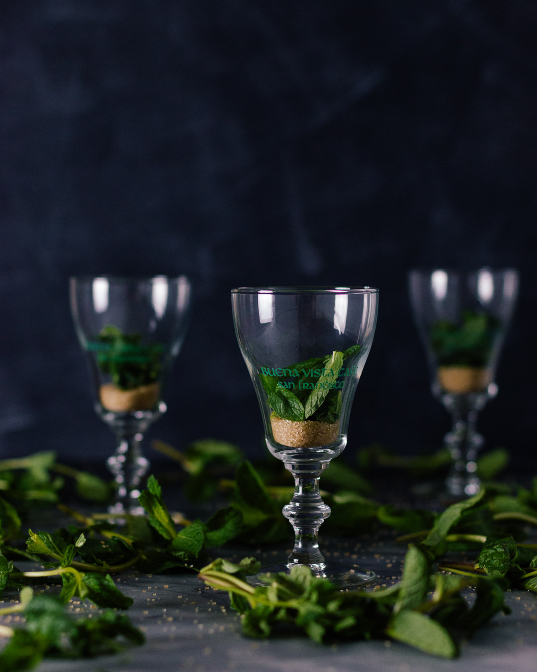 Vertical side shot of three Buena Vista Irish Coffee goblet glasses with sugar and mint leaves in them.