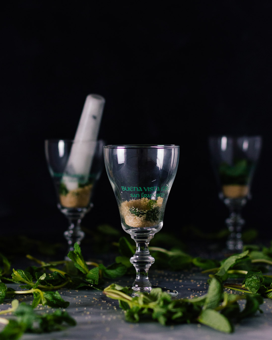 Vertical side shot of three Buena Vista Irish Coffee goblet glasses with muddled mint and sugar in them and a muddle in the back left glass.