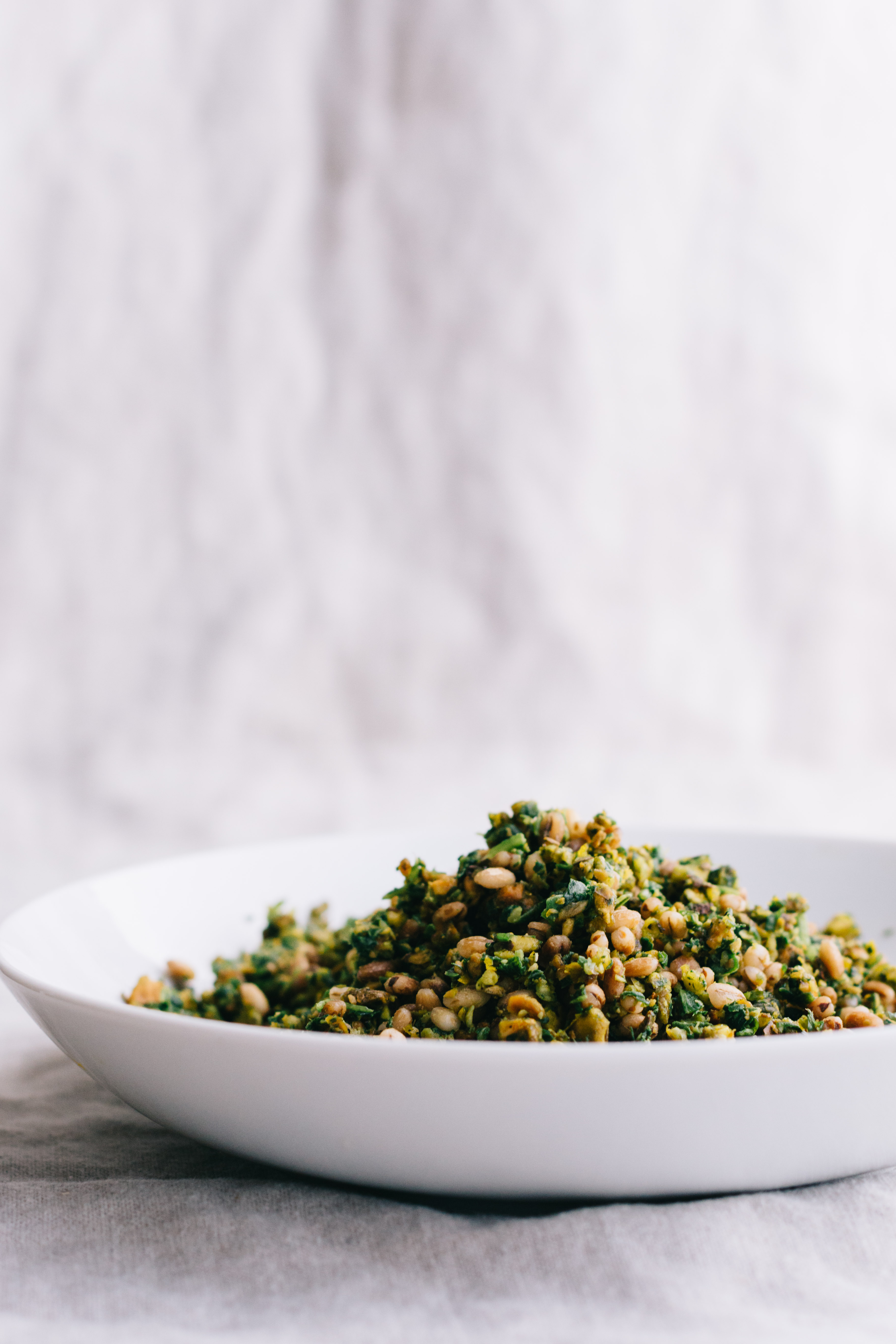 Vertical shot of pistachio gremolata with barley piled high in a shallow bowl on a cloth surface.