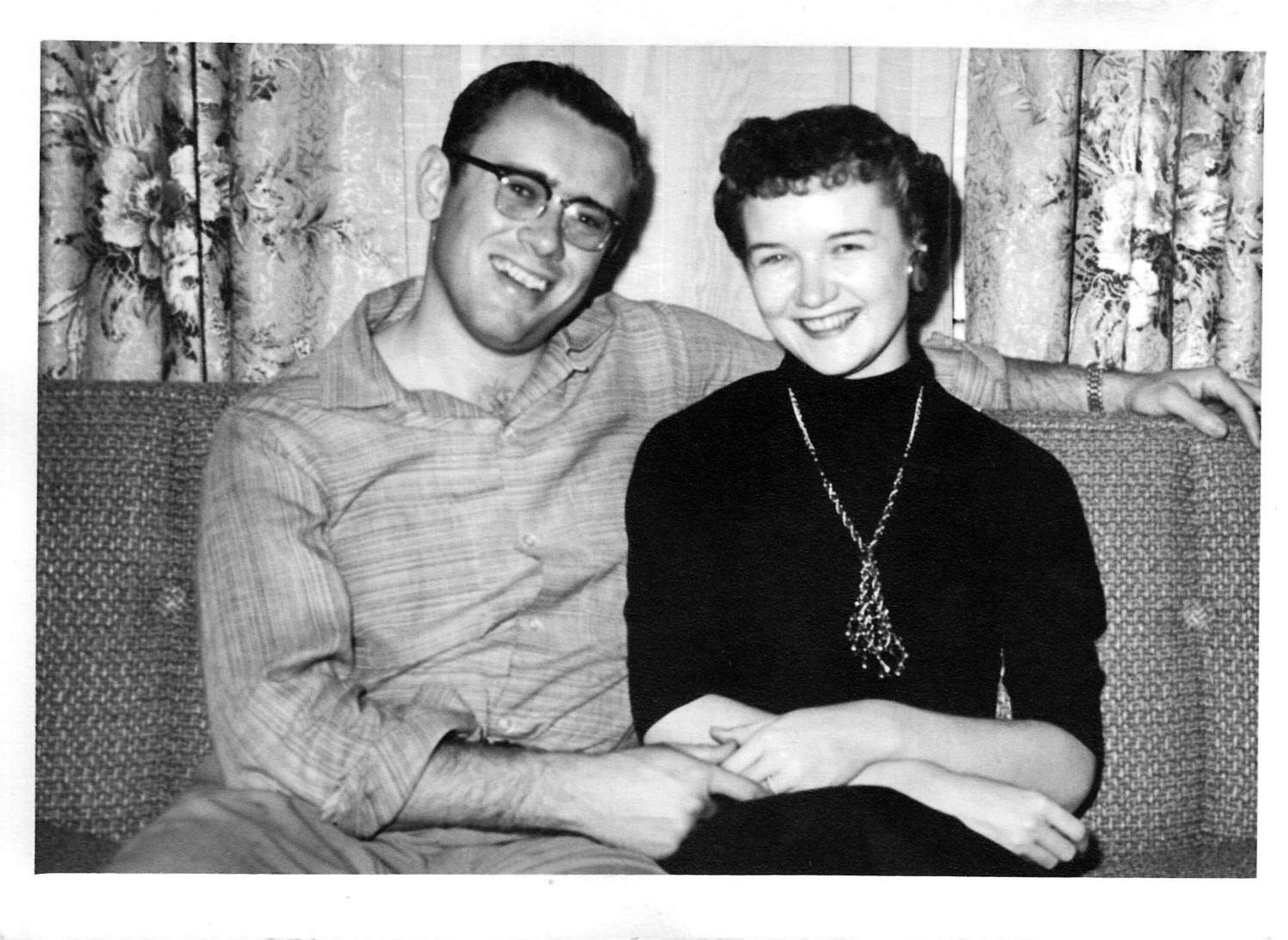 A young man with glasses and a young woman sitting on a couch next to each other and holding hands.