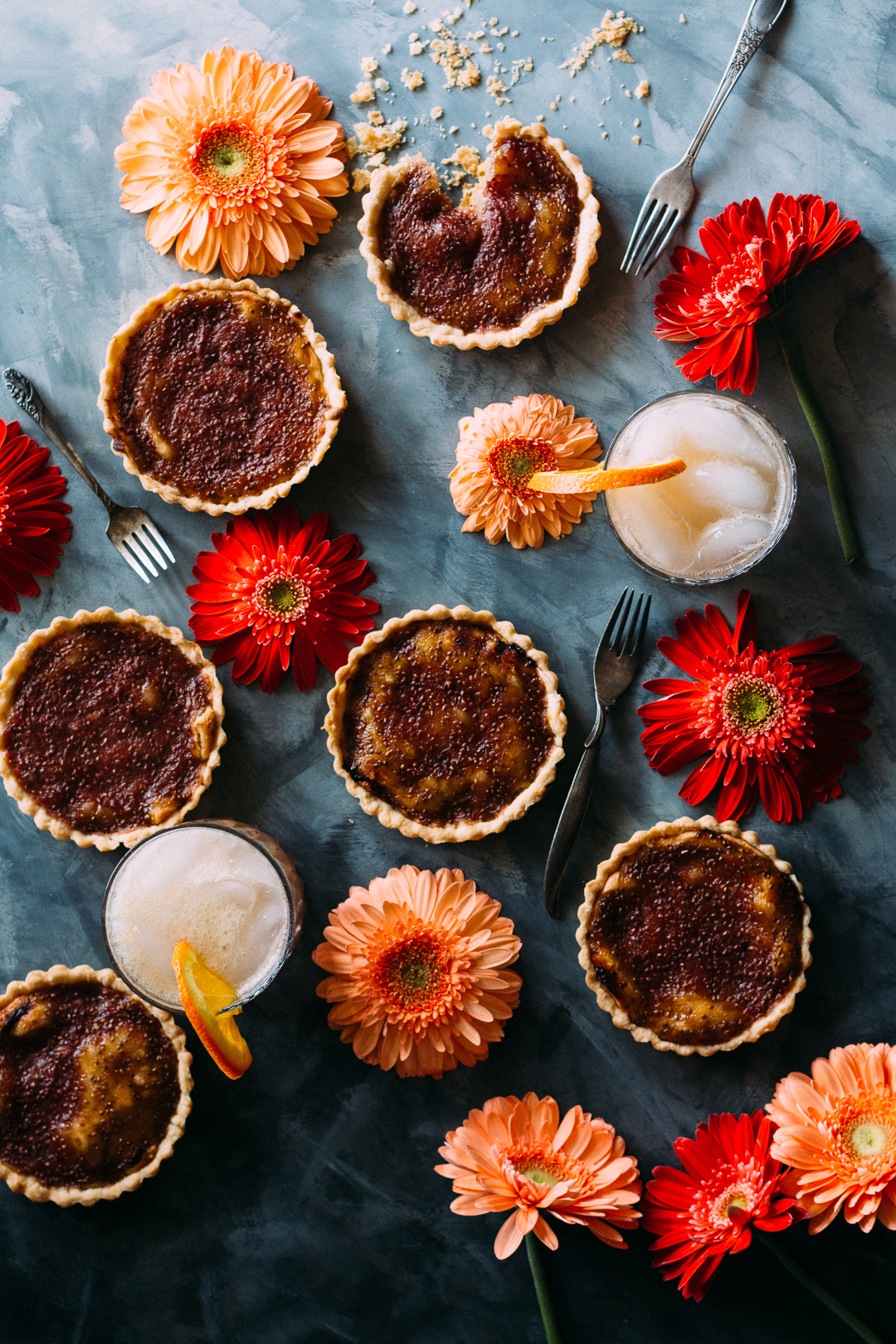 Over head shot of 6 rhubarb tarts spread out and surrounded by flowers, forks, and two drinks with orange slices in them.