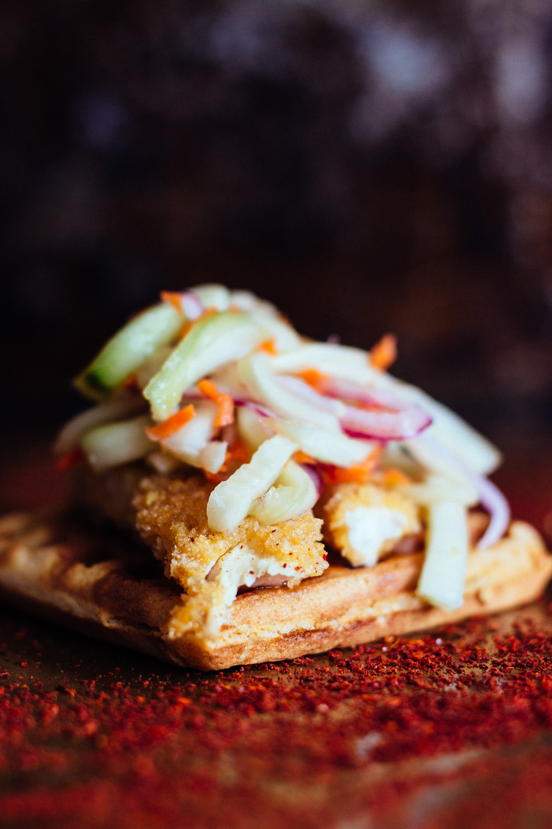 Vertical close up side shot of a waffle covered with chicken tenders and pickled veggies on top of a pepper powder covered baking sheet.