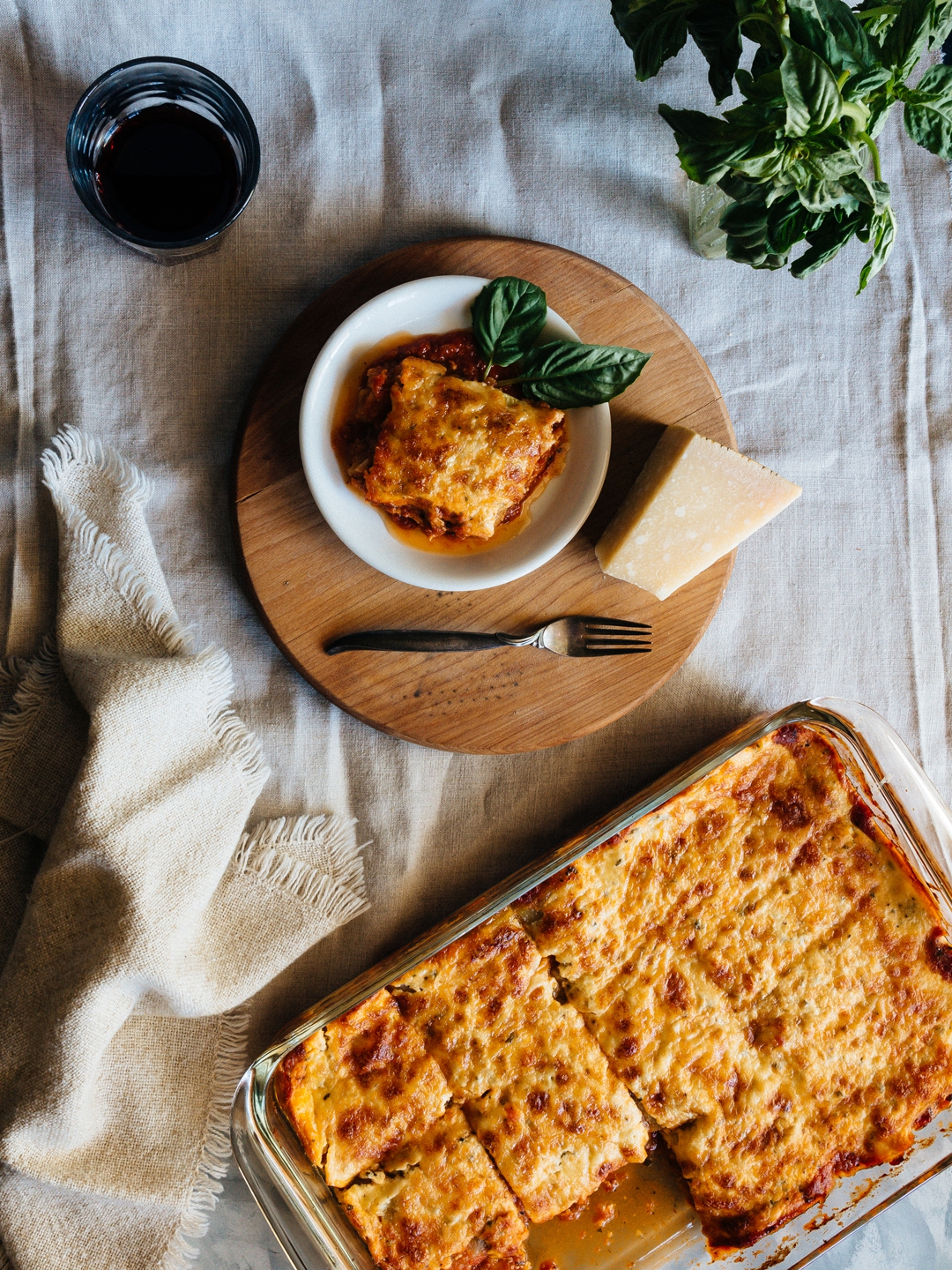 Overhead shot of a slice of zucchini lasagna on a plate surrounded by a glass of wine, basil, parmesan cheese, a fork, and a baking dish of lasagna