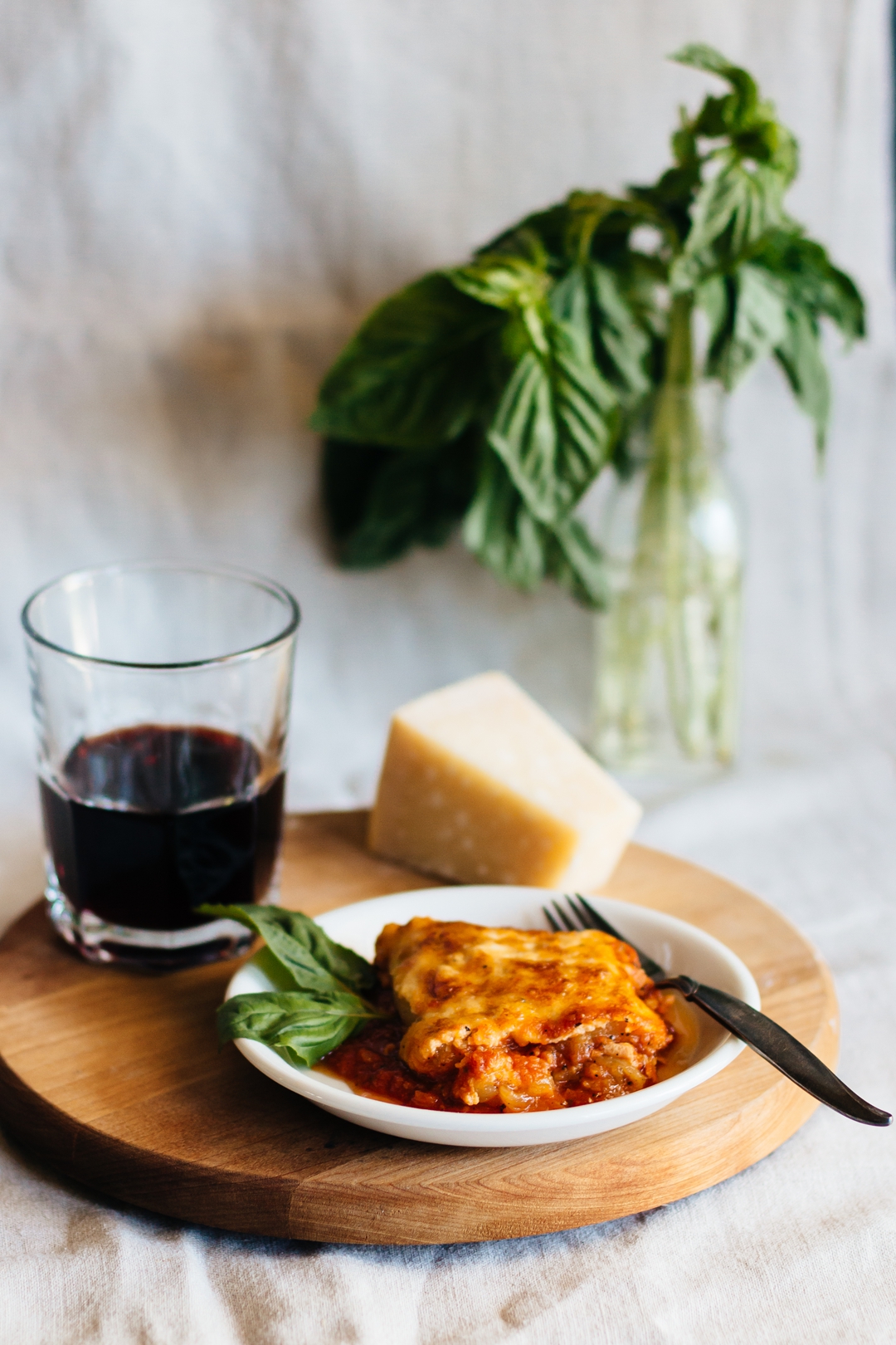 Vertical shot of a slice of zucchini lasagna on a plate with a fork surrounded by parmesan cheese and a glass of wine.