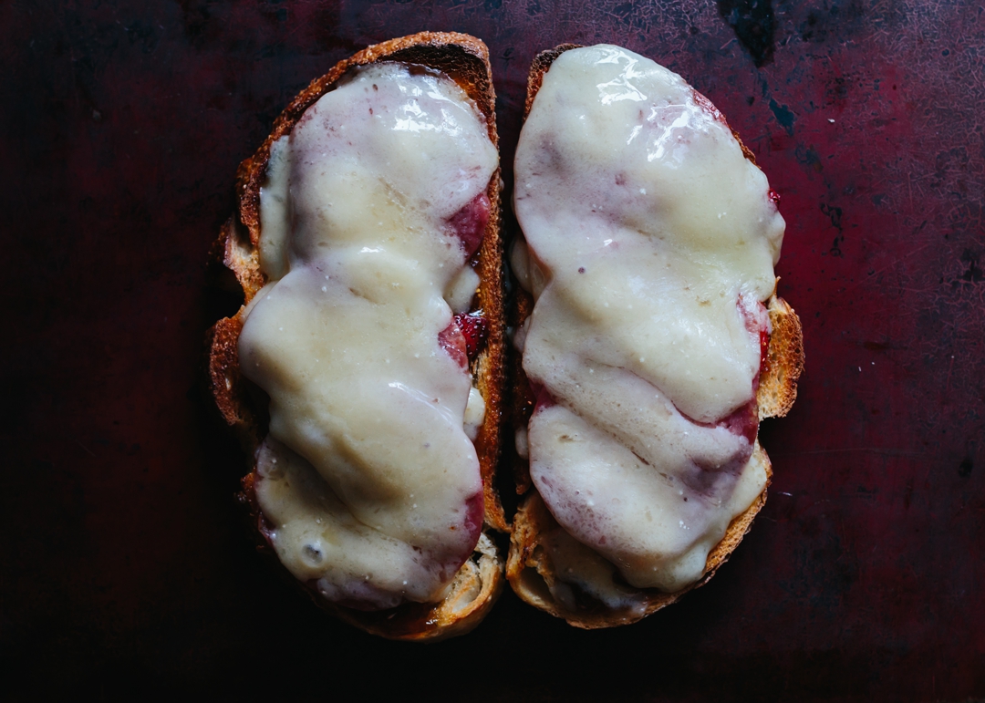Overhead shot of two slices of toast covered with slices of melted cheese, salami, strawberries, and fig jam on a baking sheet.
