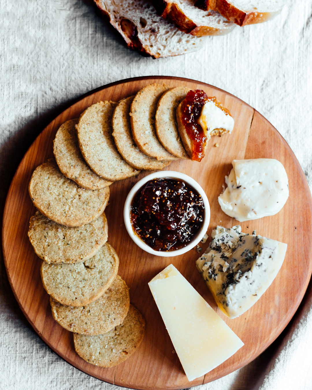 Overhead shot of pumpkin seed biscuits on round wooden cutting board with fig jam and three cheeses.