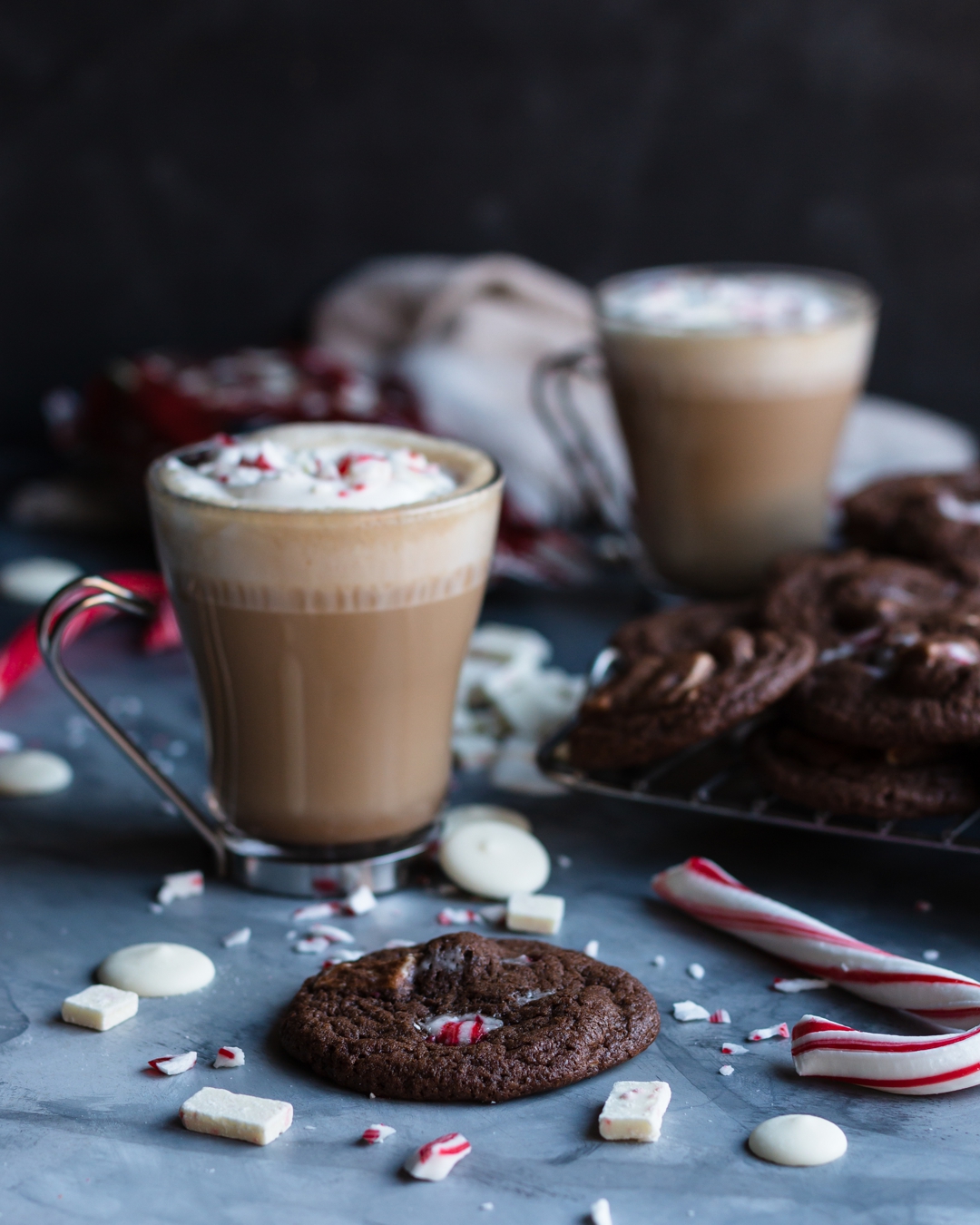 45 degrees shot of cookie surrounded by candy canes, white chocolate and peppermint chips, cookies, and two glasses with lattes in them.