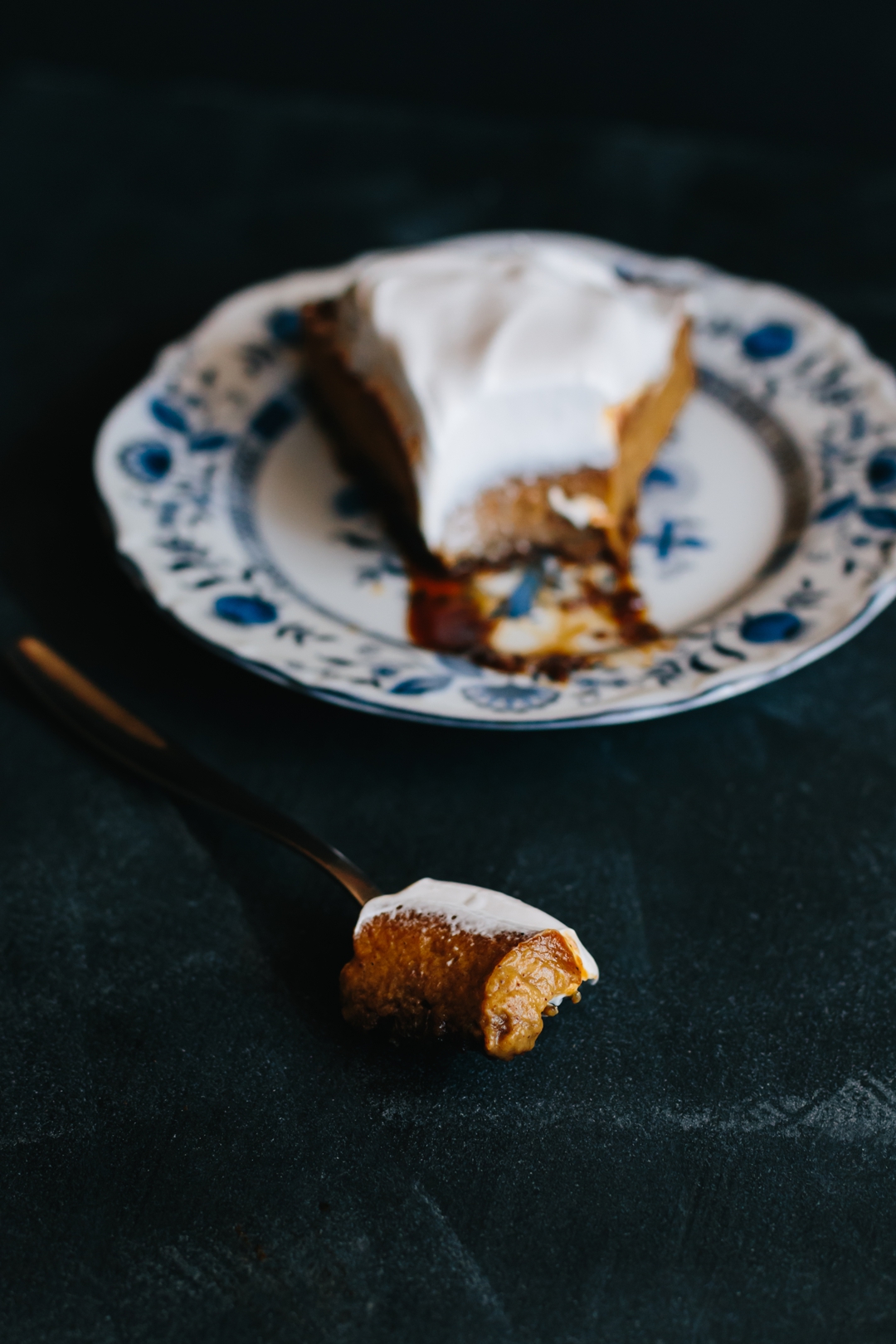 Close up shot of a bite of pumpkin pie with frosting on a fork in front of a plate with a slice on it.