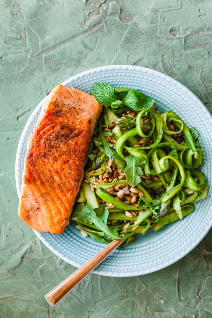 Overhead shot of a plate topped with farro, asparagus ribbons, arugula, and a salmon fillet with a piece of silver ware sticking out to the side. 