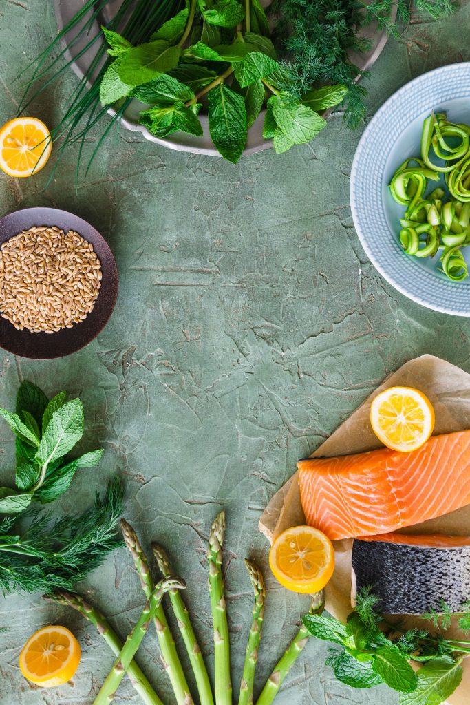 Overhead shot with empty space in the center and salmon, farro, fresh herbs, asparagus, and lemons arranged around the edges.