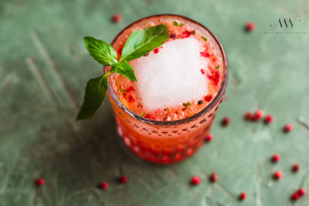 Horizontal shot of a rocks glass filled with a strawberry shrub drink, large ice cube, and basil sprig surrounded by pink peppercorns. 