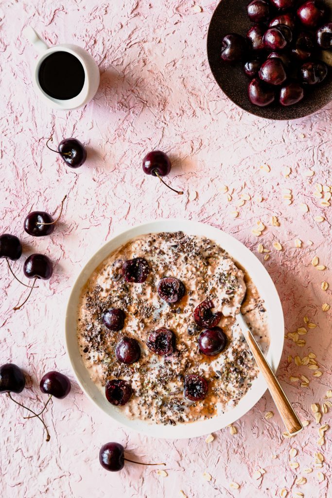 Over head shot of cherry overnight oats in a bowl with a spoon topped with cherries and surrounded by oats, cherries, and a maple syrup in a pourer. 