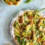Overhead shot of sautéed zucchini blossoms and herbed ricotta on a serving platter next to a bowl filled with zucchini blossoms and a smaller bowl filled with olive oil.