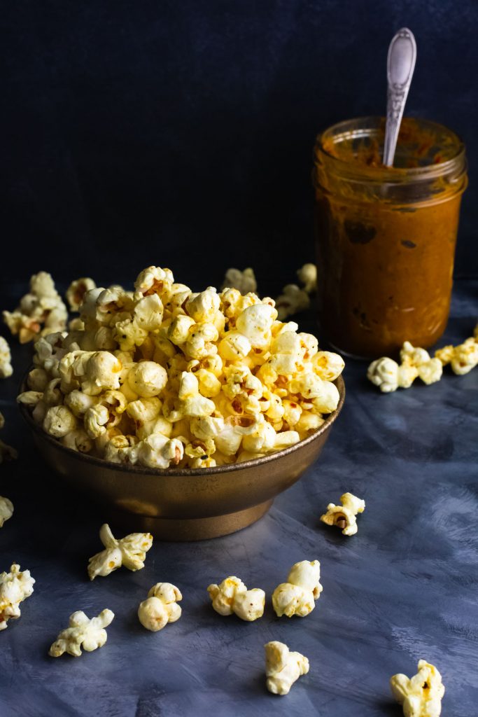 Vertical Shot of popcorn overflowing from a bowl with a mason jar of curry in the background.