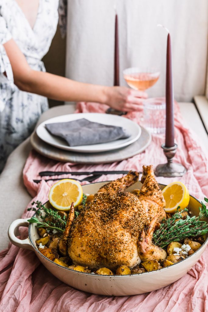 Side shot of roasted chicken in skillet on a table with candles, table setting and woman reaching for glass filled with wine.