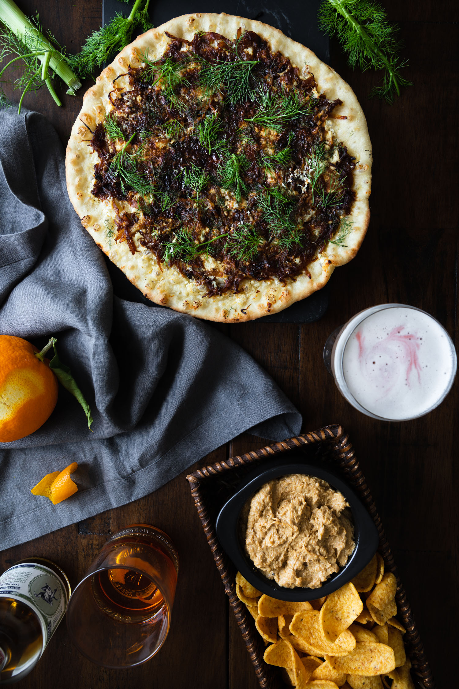 Overhead shot of Caramelized Fennel & Onion Skillet Pizza Recipe surrounded by a cloth napkin, fennel fronds, an orange, drinks, and onion dip.