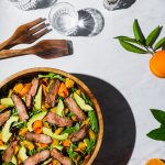Overhead shot of a flank steak salad in a wooden bowl surrounded by oranges, avocado, and glasses of water.